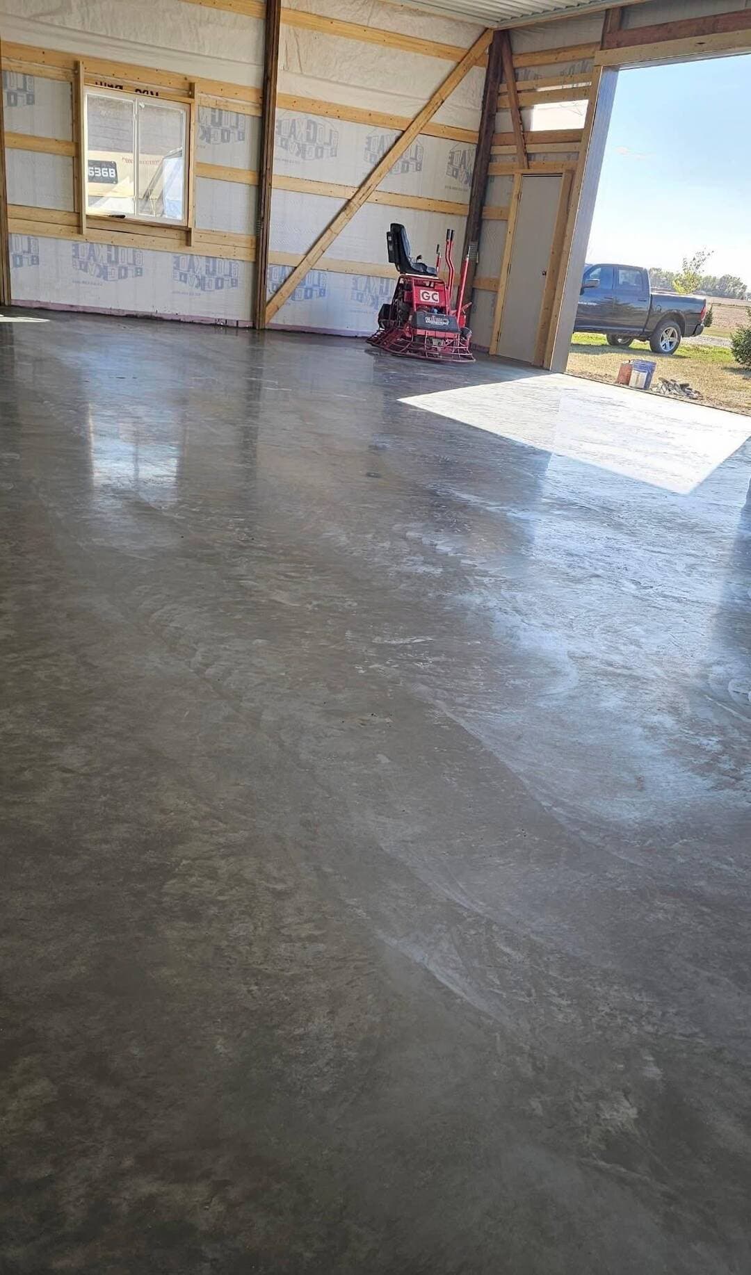 A freshly sealed pole-barn floor shining inside the framed building, trowel machine at the door