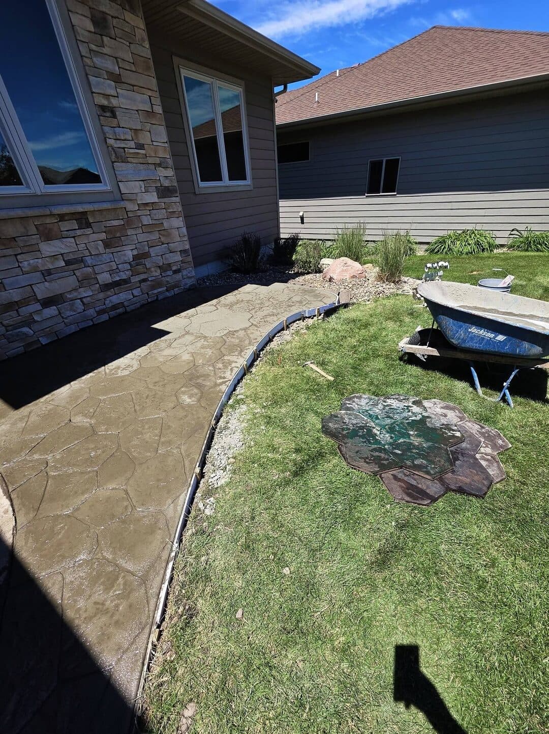 A freshly stamped flagstone walkway curving behind a stone home, stamp mats and a wheelbarrow still on the lawn
