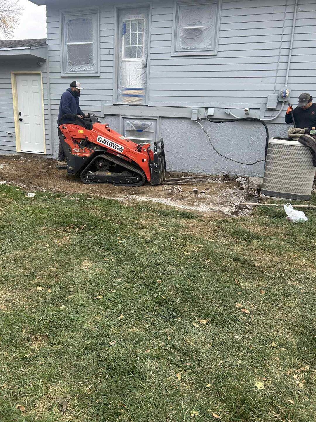 A tracked skid steer grading along the side of a house